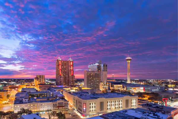 City skyline at sunset with a purple-pink sky, tall illuminated towers, and snow-covered streets.