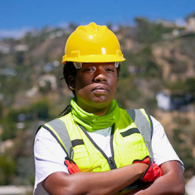 worker-1jpeg - San Antonio Gold Buyers - San Antonio Gold Buyers Construction worker wearing a yellow hard hat and high-visibility vest with arms crossed at a construction site.