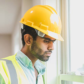 worker-3jpeg - San Antonio Gold Buyers - San Antonio Gold Buyers Construction worker wearing a yellow hard hat and reflective vest, looking out a window.