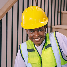 worker-4jpeg - San Antonio Gold Buyers - San Antonio Gold Buyers Smiling construction worker in a yellow hard hat and neon safety vest indoors near a staircase.